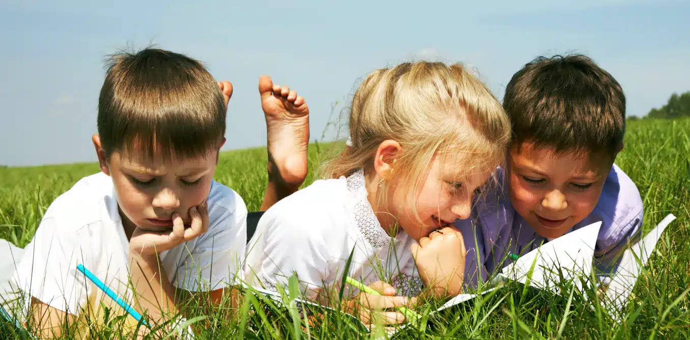 Three children lying in the grass, engrossed in a book. Outdoor learning enhances child development