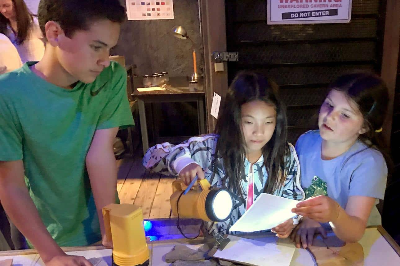 Three children studying a paper on a table in an Escape Room, following safety and provision guidelines