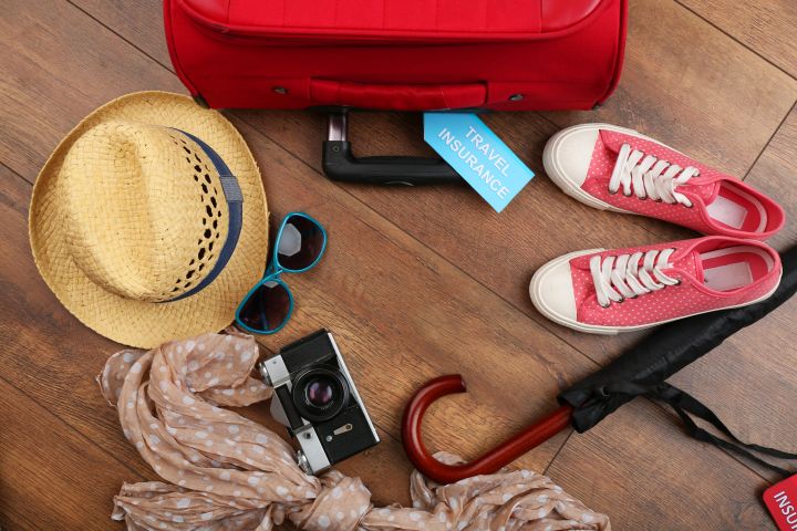 A red suitcase, hat, sunglasses, camera, and more on a wooden floor - essentials for a budget-friendly holiday