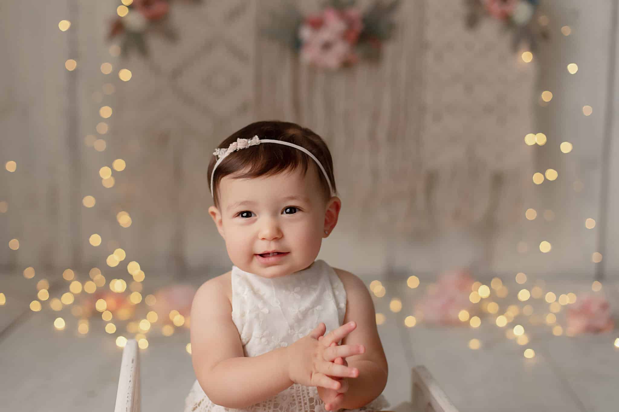 A baby girl sitting in a wooden chair with string lights illuminating the background