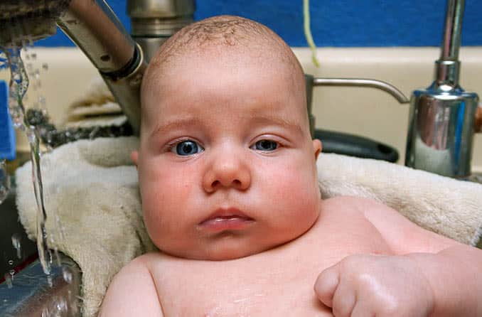 A baby sitting in a sink with water running, showcasing Eyelid Drooping - Ptosis