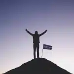 A photograph of a lone figure in silhouette with arms raised triumphantly atop a stark,