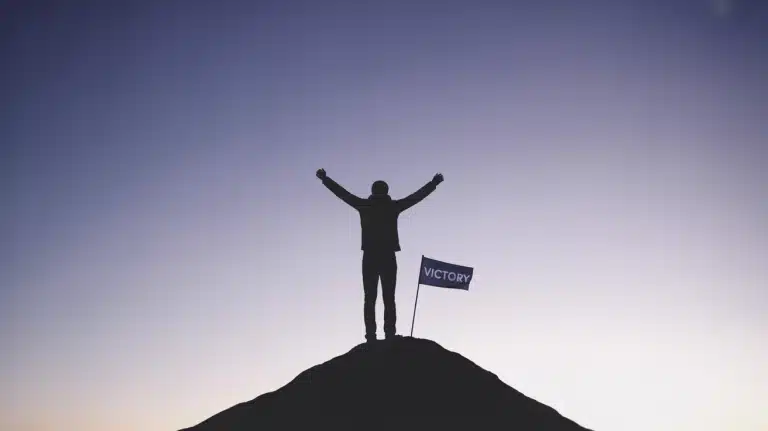 A photograph of a lone figure in silhouette with arms raised triumphantly atop a stark,